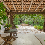 A covered patio featuring a stone path leading to a table, surrounded by greenery and outdoor decor